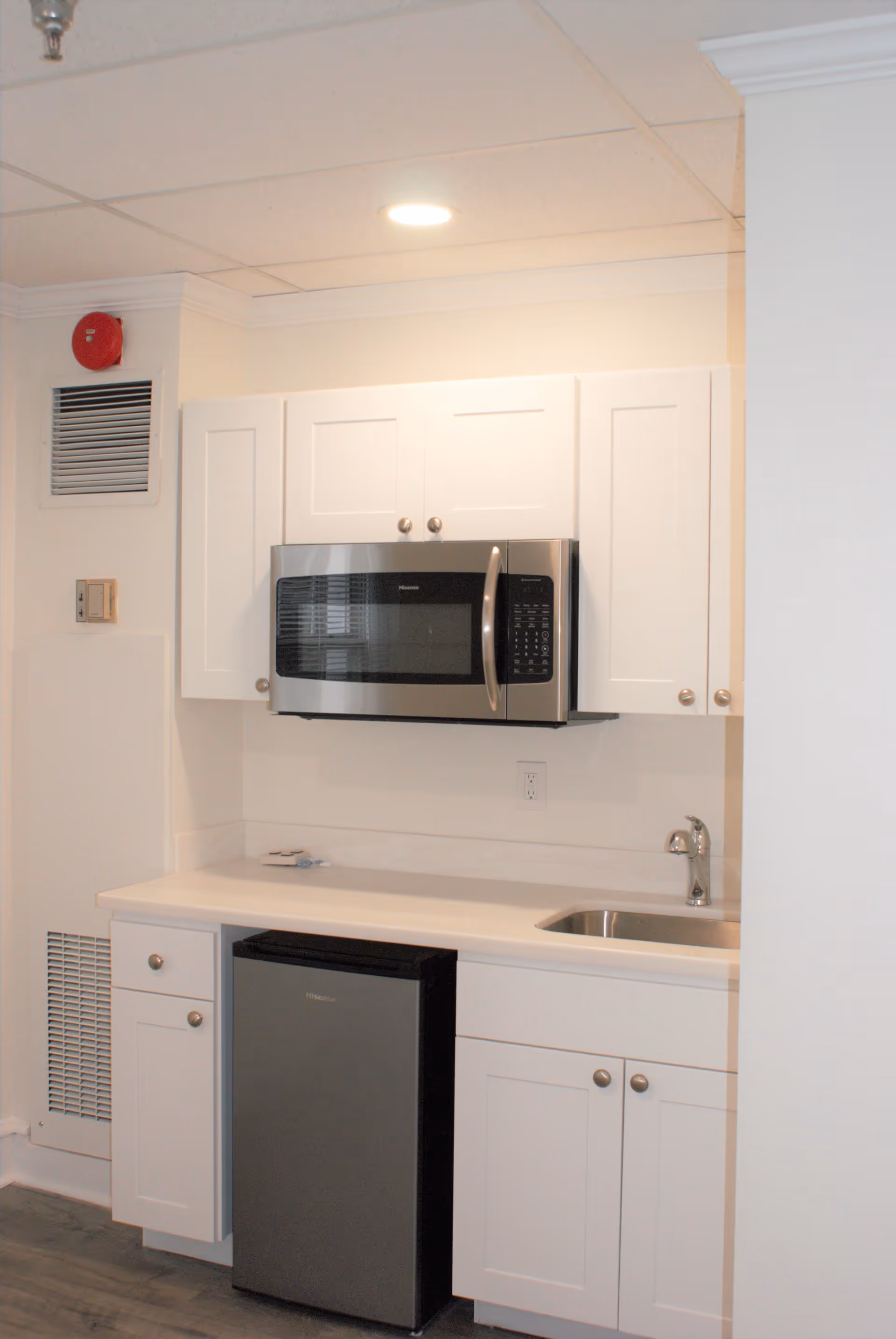 A small kitchenette with white cabinets, a stainless steel microwave mounted above the counter, a small refrigerator below the counter, and a sink with a faucet on the right side. The walls and ceiling are white, and there is a recessed light above the microwave.