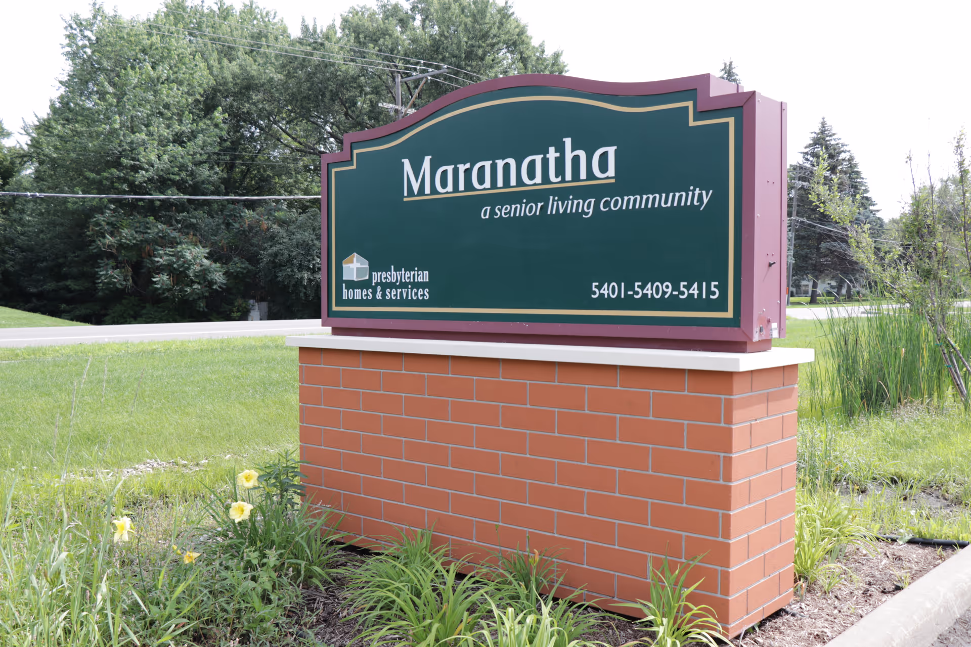 Outdoor image of a green and maroon sign for Maranatha, a senior living community, mounted on a brick base with grass, flowers, and trees in the background.
