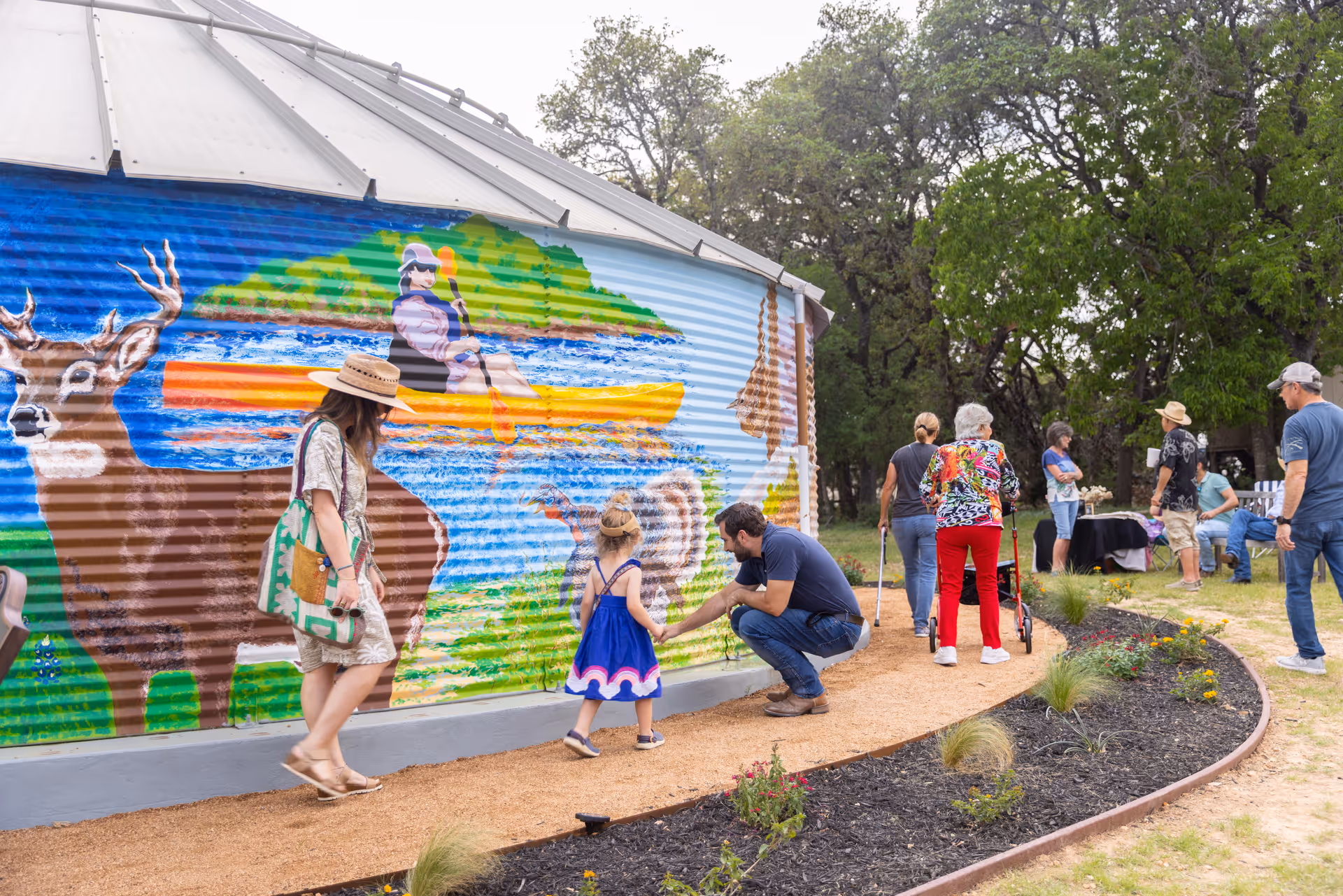 A group of people, including a man crouching to talk to a young girl in a blue dress, walk along a curved path next to a large colorful mural depicting a deer and a person kayaking. The scene is outdoors with trees and a small garden bed along the path.