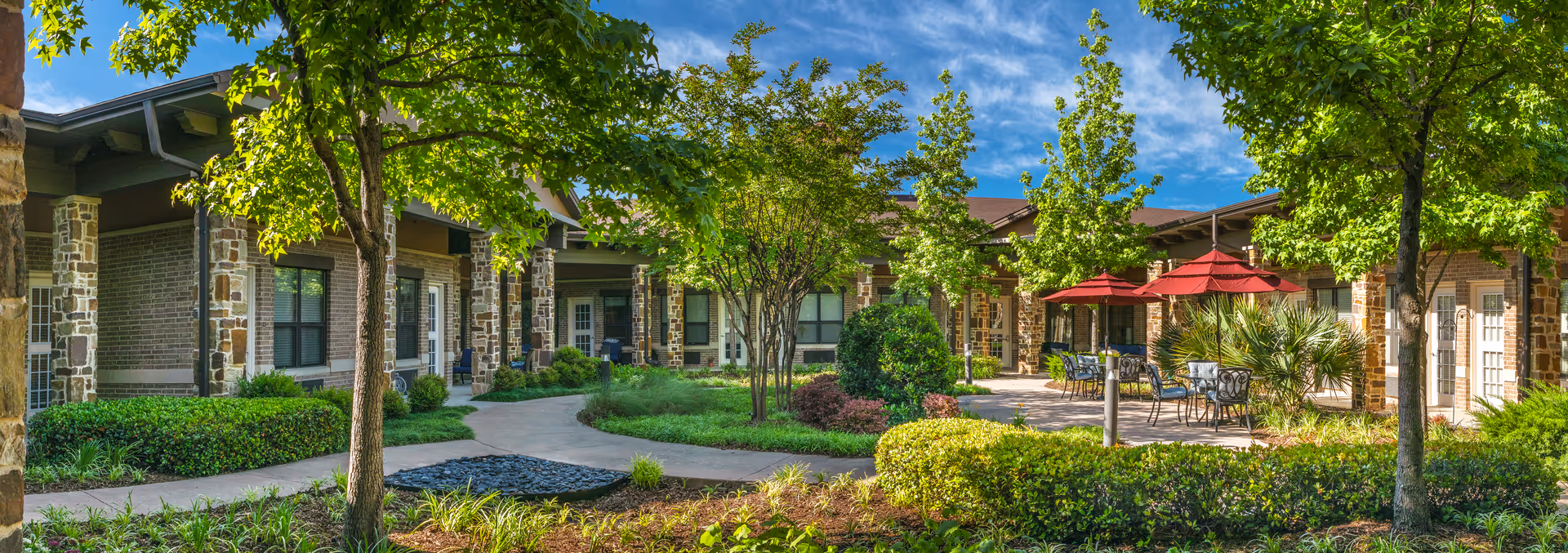 Outdoor courtyard area at The Villages of Lake Highlands featuring a paved walkway, green trees, shrubs, and patio tables with red umbrellas under a blue sky.