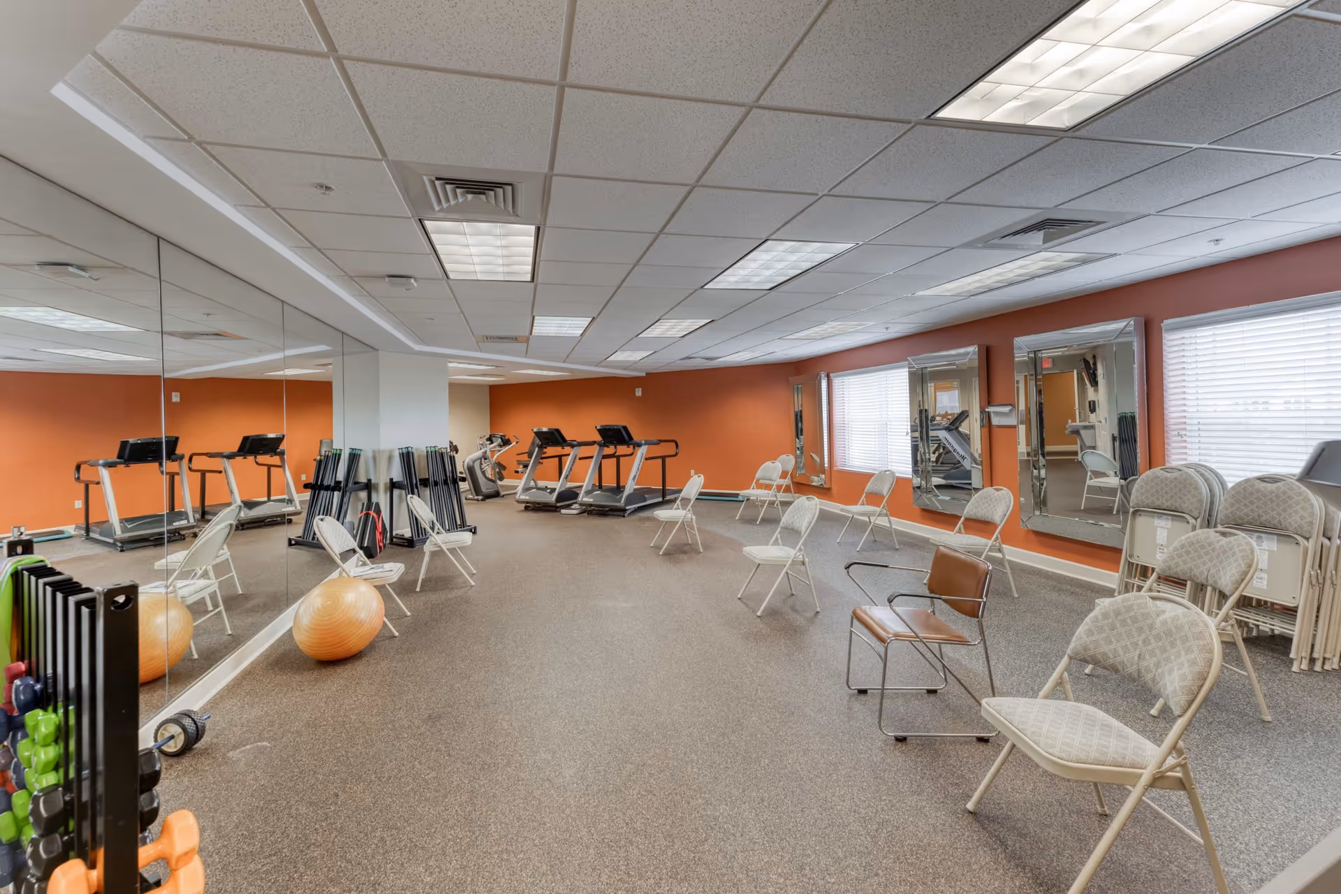 Exercise room with treadmills, folding chairs, mirrors, and fitness equipment in a senior living facility.