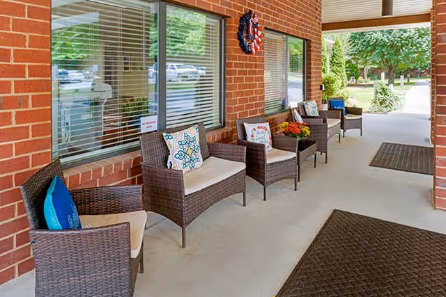 Covered outdoor seating area with several wicker chairs featuring cushions and decorative pillows, placed along a brick wall with windows. There are two dark floor mats on the concrete floor and greenery visible in the background.