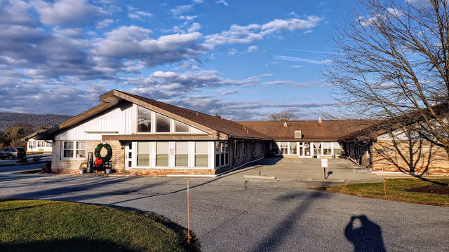 Exterior view of a single-story senior living facility building with a sloped roof, large windows, and a wreath decoration near the entrance. The sky is partly cloudy and there is a paved driveway and some grass areas around the building.