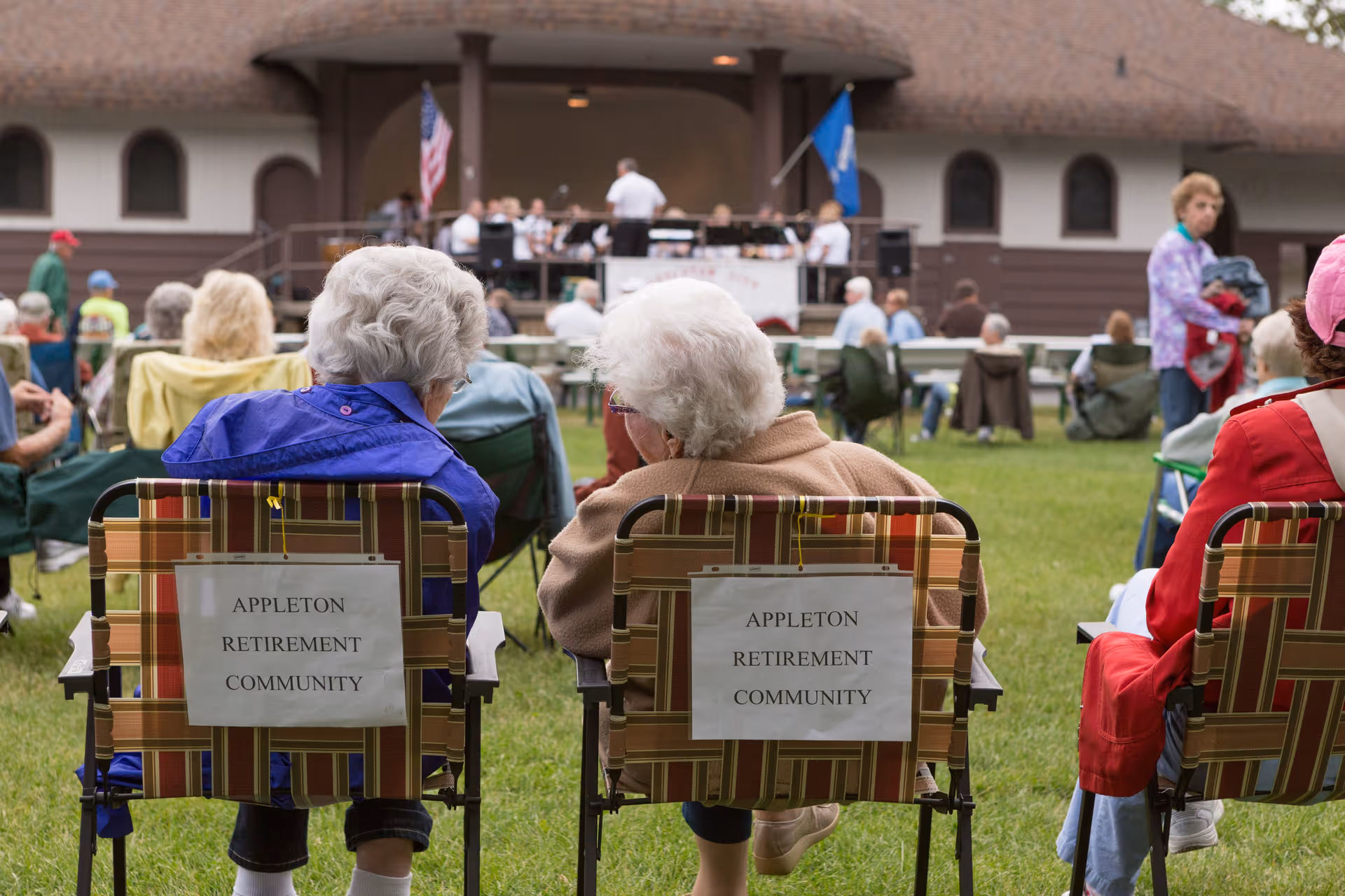 Two elderly women sitting on lawn chairs with signs reading 'Appleton Retirement Community' on the back, watching a band performing on an outdoor stage with American and state flags.