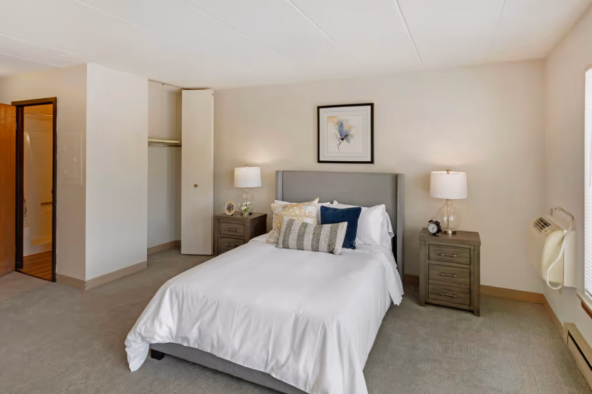A neatly made bed with white bedding and multiple pillows in a senior living bedroom. The room features two wooden nightstands with lamps on each side of the bed, a framed artwork above the headboard, an open closet with a sliding door, and a window with blinds. There is a door leading to a bathroom on the left side and a wall-mounted air conditioning unit on the right.