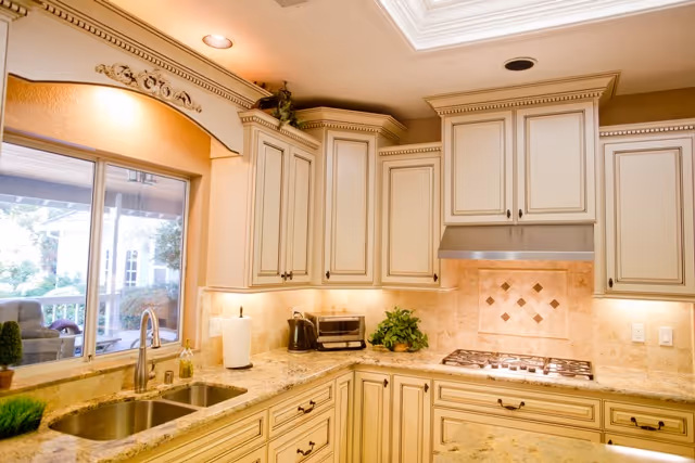 A bright and elegant kitchen with cream-colored cabinets, granite countertops, a double sink under a large window, a built-in gas stove with a stainless steel range hood, and small kitchen appliances like a toaster and a coffee maker. There is a decorative tile backsplash and recessed lighting in the ceiling.