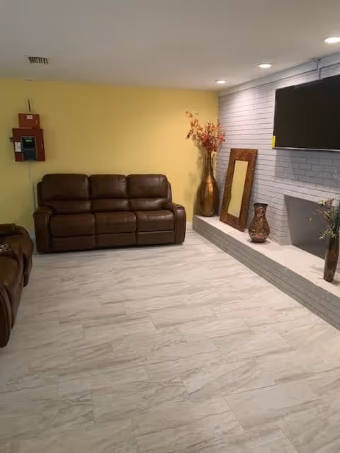 A living room with a brown leather sofa against a yellow wall, a white brick fireplace with decorative vases and a large mirror resting on the hearth, and a flat-screen TV mounted above the fireplace. The floor is light-colored wood or tile, and the room is lit with recessed ceiling lights.