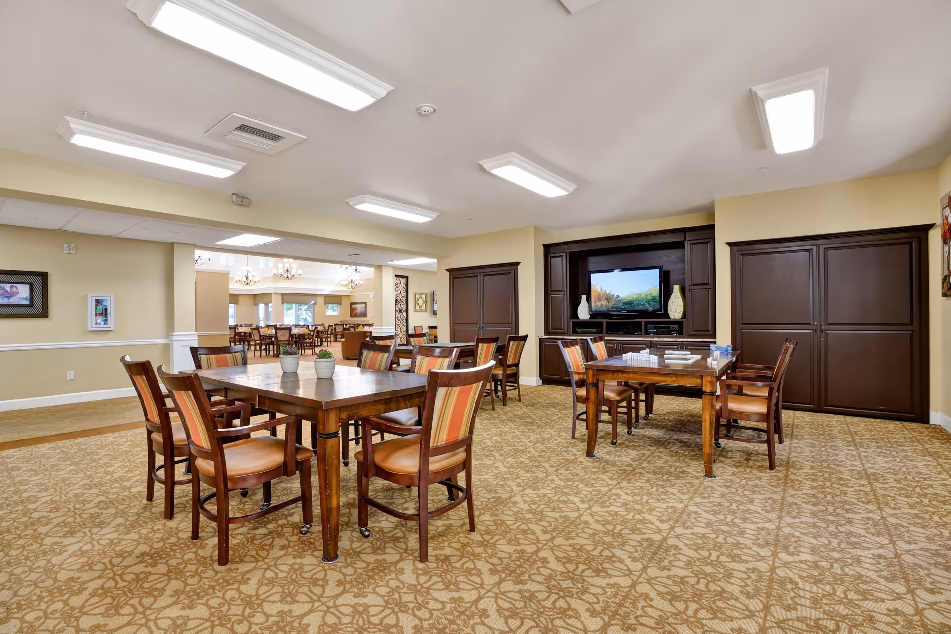 A spacious common area with multiple wooden tables and chairs arranged on a patterned carpet. The room has beige walls and ceiling lights. In the background, there is a large flat-screen TV mounted within a dark wood entertainment center, flanked by decorative vases. Beyond this room, another dining area with more tables and chairs is visible.