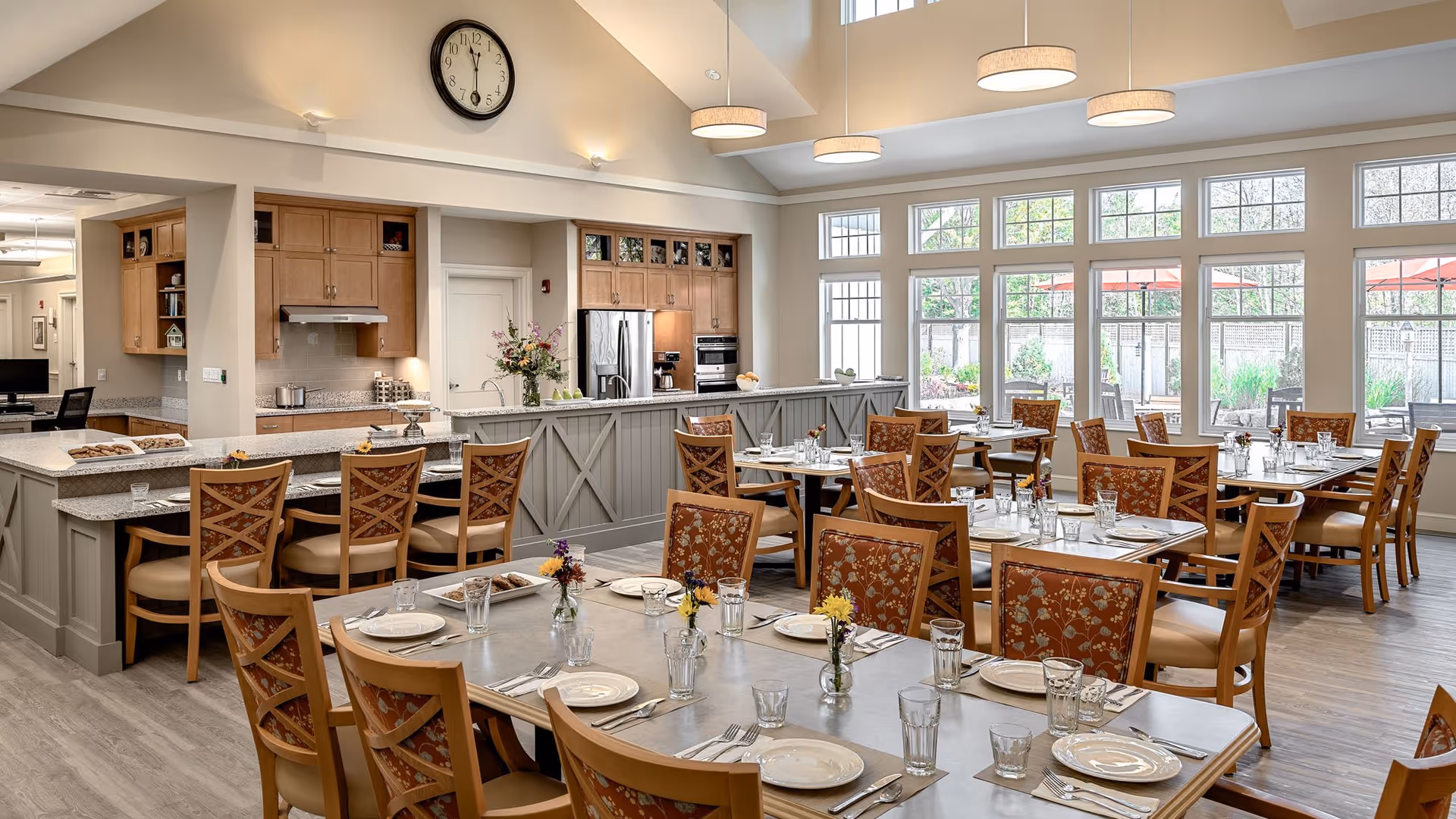 A bright and spacious dining room with multiple tables set with plates, glasses, and silverware. The chairs have wooden frames with floral upholstery. The room features large windows letting in natural light and a view of an outdoor patio area with umbrellas. In the background, there is a kitchen area with wooden cabinets, a refrigerator, and a clock on the wall.