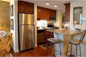 Open kitchen with stainless steel refrigerator, wooden cabinets, a granite breakfast bar with two barstools, and part of the adjacent living area visible.