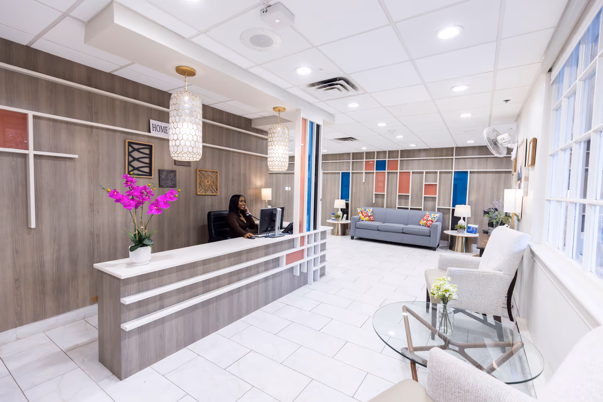 Bright reception lobby with a front desk and receptionist on the left, a seating area with a sofa in the background, and chairs with a glass table by the window on the right.