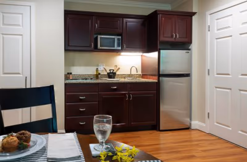 A small kitchen area with dark wood cabinets, a granite countertop, a stainless steel refrigerator, a microwave, and a sink. In the foreground, part of a dining table is visible with a glass of water, a plate with muffins, a napkin, and a yellow flower on the table. The floor is wooden, and there are two white doors on either side of the kitchen.