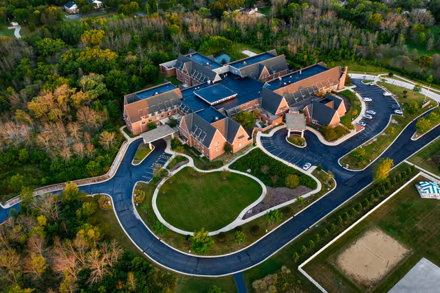 Aerial view of the Teal Shores senior living complex with a central brick building, circular drive, landscaped lawns, and surrounding trees.