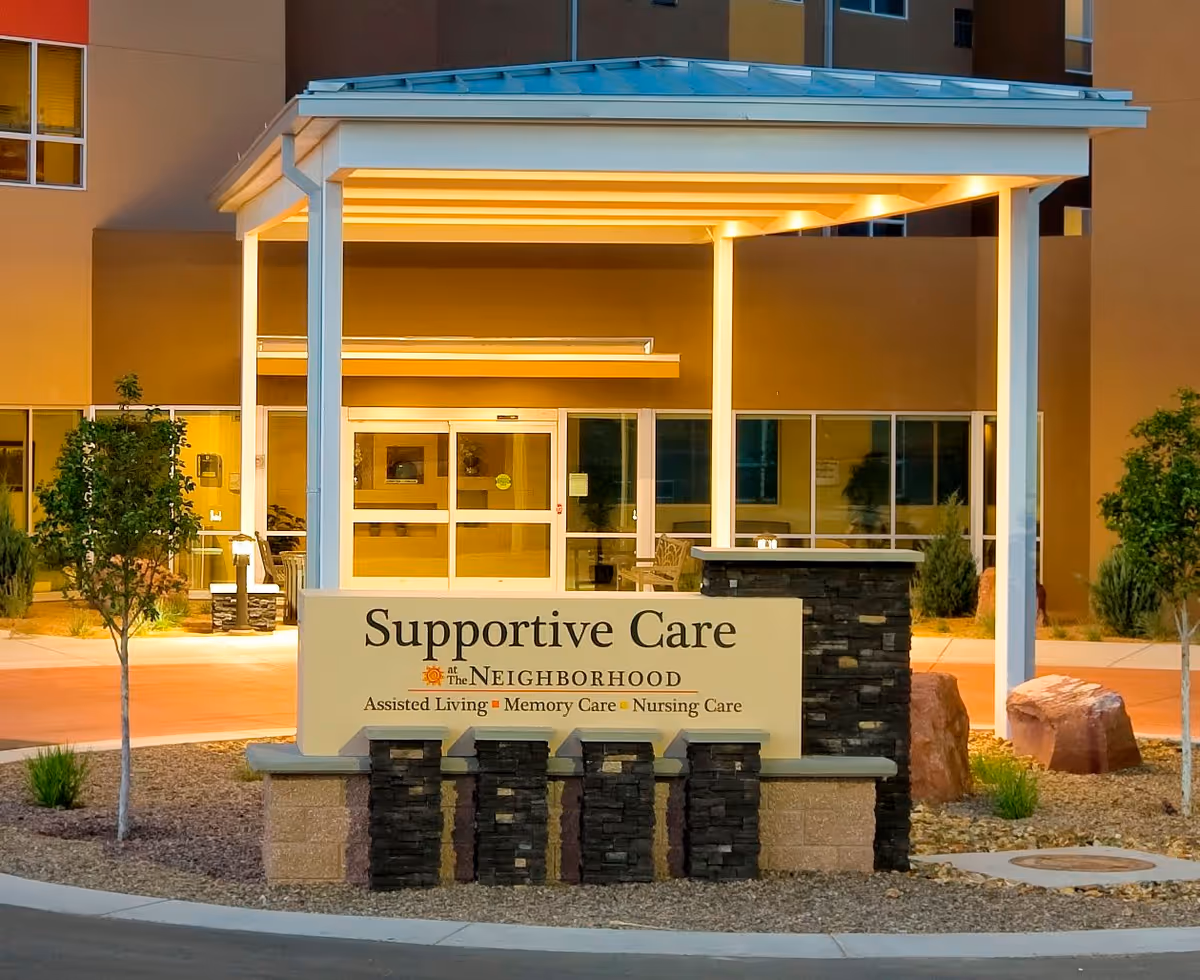 Entrance to the Supportive Care section of The Neighborhood in Rio Rancho Life Plan Community, featuring a covered walkway, illuminated sign, and landscaped area with small trees and rocks.