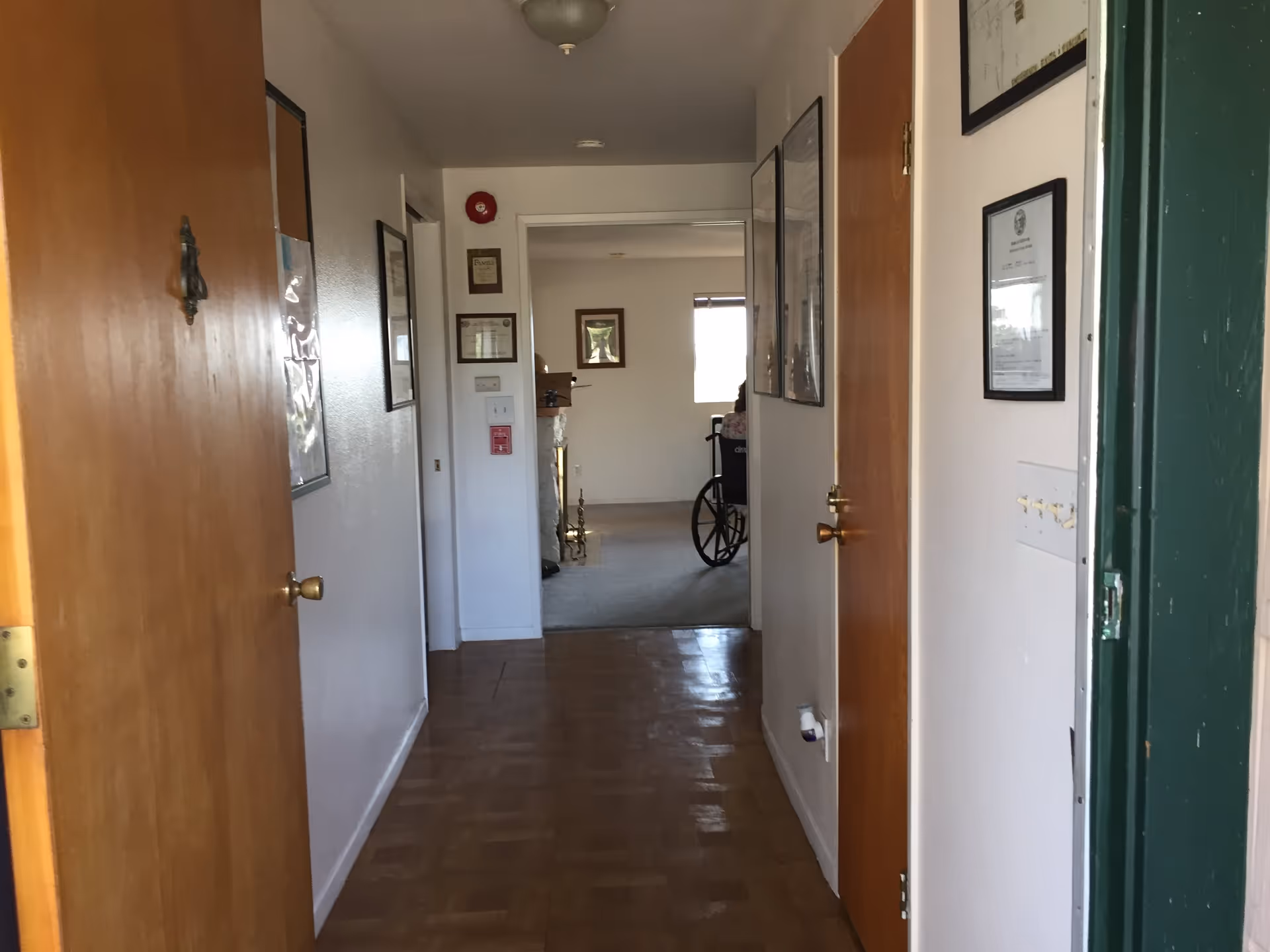 View down a hallway in a senior living facility with wooden doors on both sides and framed certificates and pictures on the walls. At the end of the hallway, there is a room with a window and a person sitting in a wheelchair.