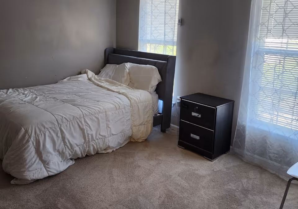 Simple bedroom with a made single bed against a wall, a black two-drawer nightstand, and light filtering through windows with blinds and sheer curtains.