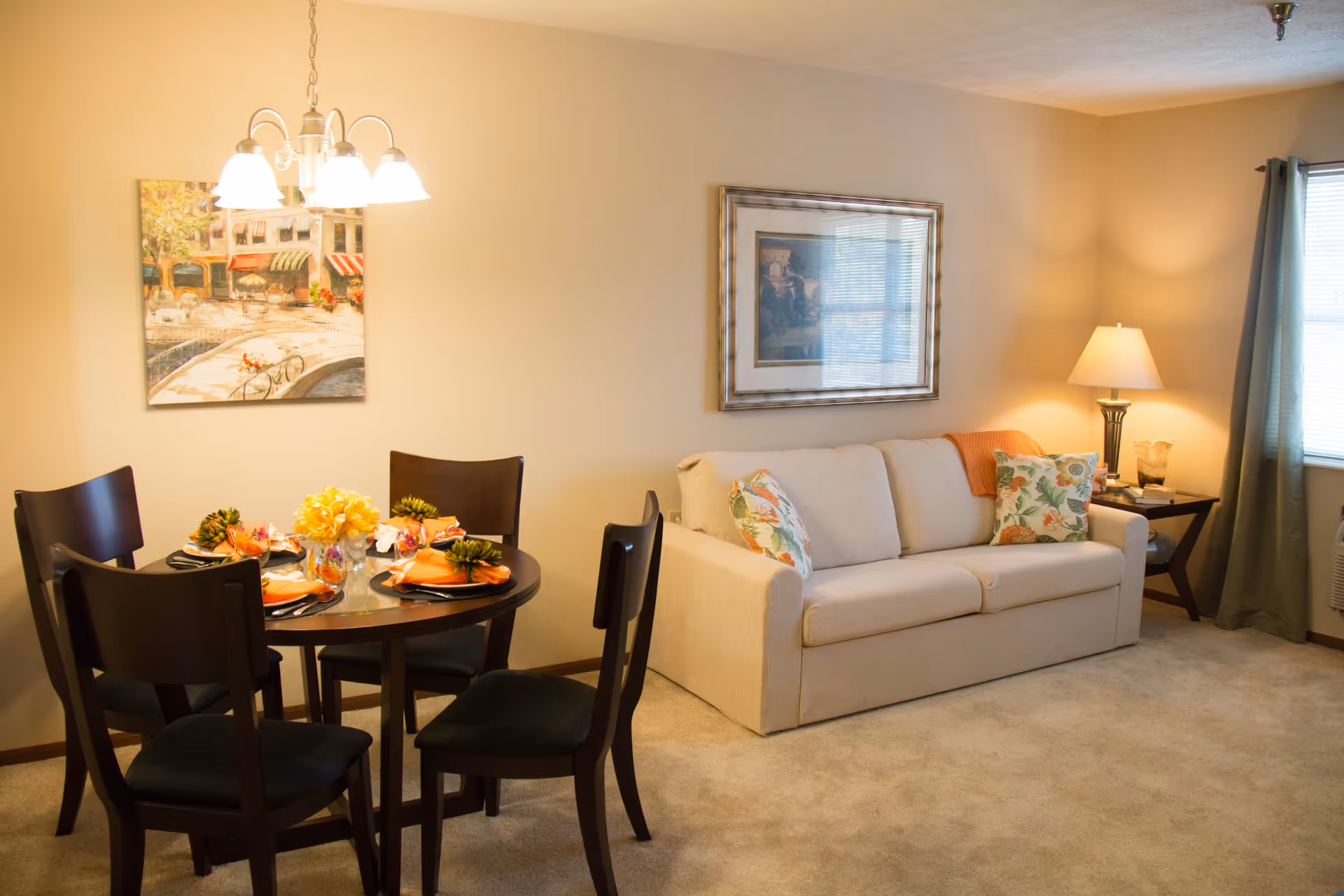 A cozy living and dining area with a beige sofa adorned with floral pillows and an orange throw blanket. Next to the sofa is a side table with a lamp and decorative items. A round dining table set for four with dark wooden chairs is positioned under a chandelier. The walls are decorated with framed artwork, and a window with green curtains allows natural light into the room.