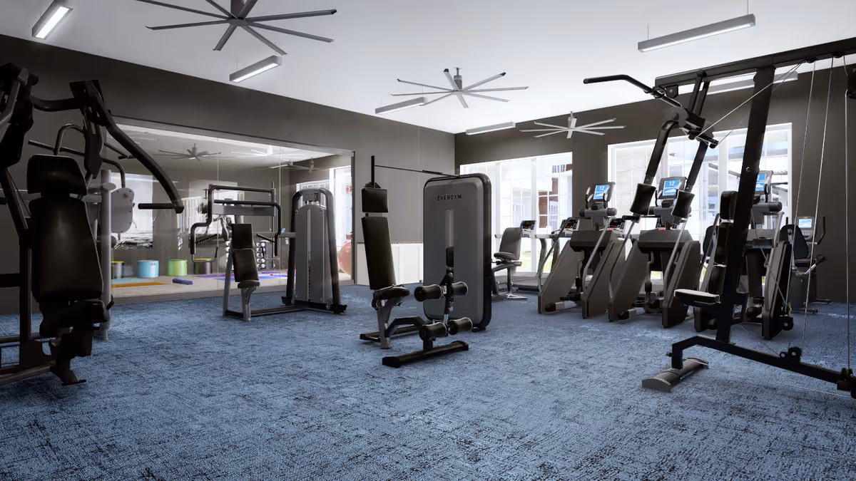 A modern fitness room with various exercise machines including weight machines and cardio equipment. The room has large windows letting in natural light, a blue carpeted floor, and ceiling fans for ventilation. There is a large mirror on one wall reflecting the equipment.