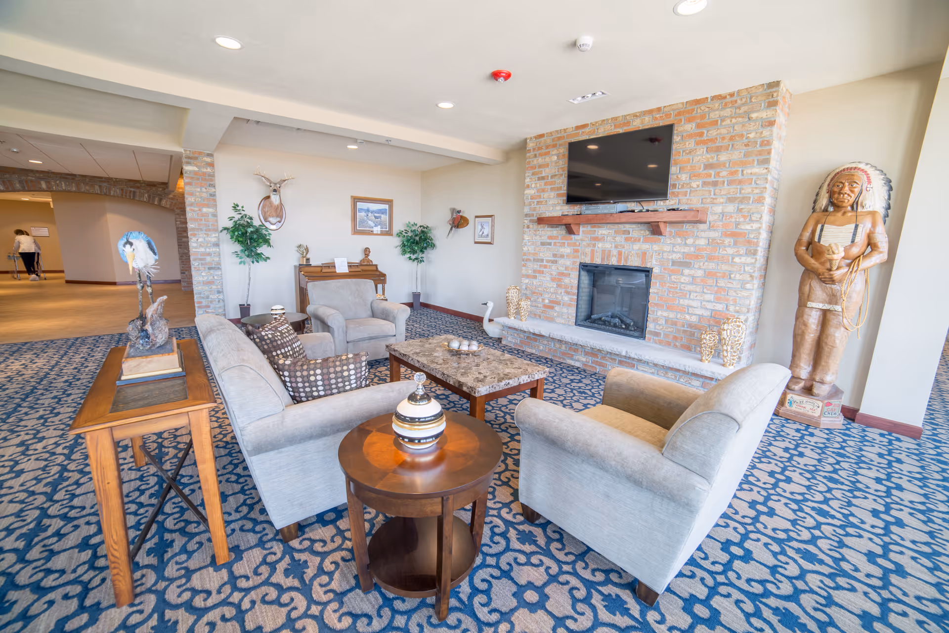 A cozy living room area in an assisted living facility featuring a brick fireplace with a mounted flat-screen TV above it. The room has a patterned blue carpet, two armchairs, a sofa with patterned cushions, a wooden coffee table, and a side table with a decorative vase. There are two potted plants, a mounted deer head, framed pictures on the wall, and a wooden statue of a Native American figure standing near the window.