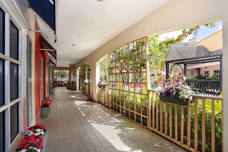 A bright covered outdoor walkway with tiled flooring and a wooden trellis on one side adorned with flowering plants. The other side features windows and red walls with flower pots placed along the base. In the background, there are more plants and a seating area under a pergola.