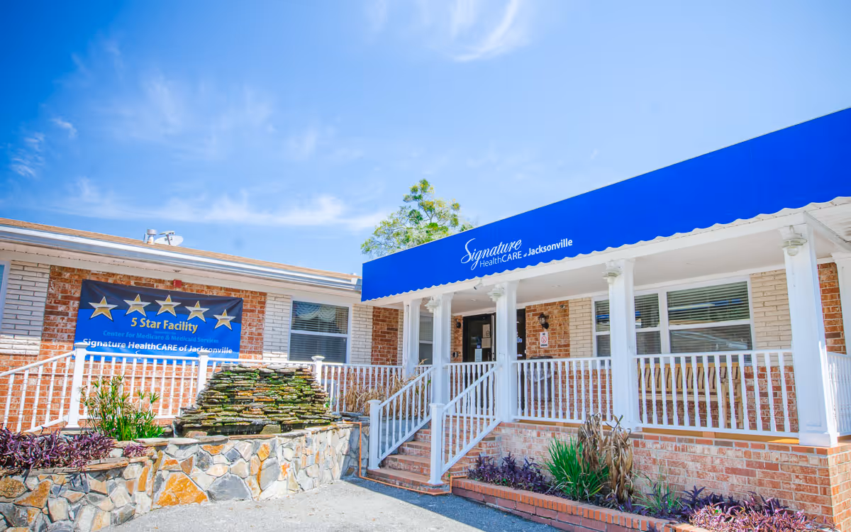 Front entrance of a brick nursing and rehabilitation facility with a blue awning, white porch railings, and a five-star banner.