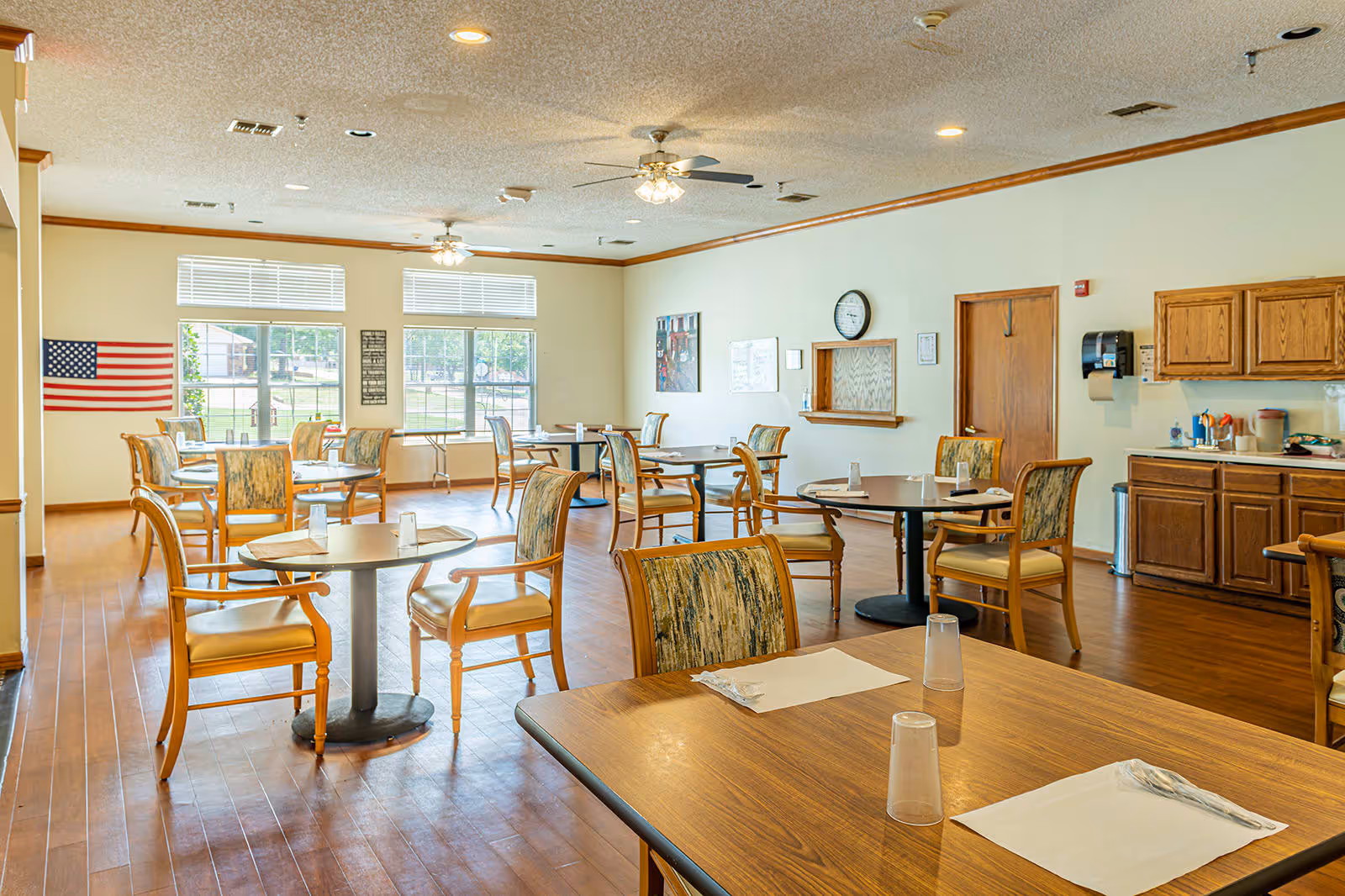 A bright dining room with multiple round tables and wooden chairs with cushioned seats and backs. The tables are set with placemats, plastic cups, and utensils. Large windows allow natural light to fill the room. An American flag is displayed on the wall, and there is a clock and a serving window on another wall. Wooden cabinets and a countertop with various items are visible on the right side.