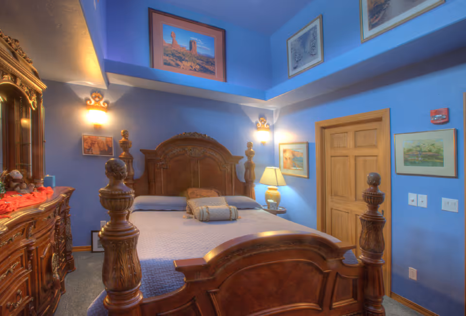 Ornate wooden bed and dresser in a blue-painted bedroom with framed artwork and bedside lamps.