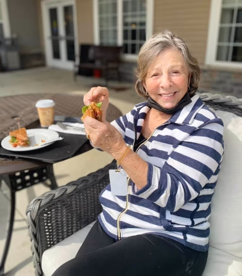 An elderly woman wearing a striped jacket and a black face mask under her chin is sitting on a cushioned outdoor chair, smiling and holding a sandwich. On the round table in front of her, there is a plate with a sandwich and a cup of coffee. The setting appears to be an outdoor patio area with a building and windows in the background.