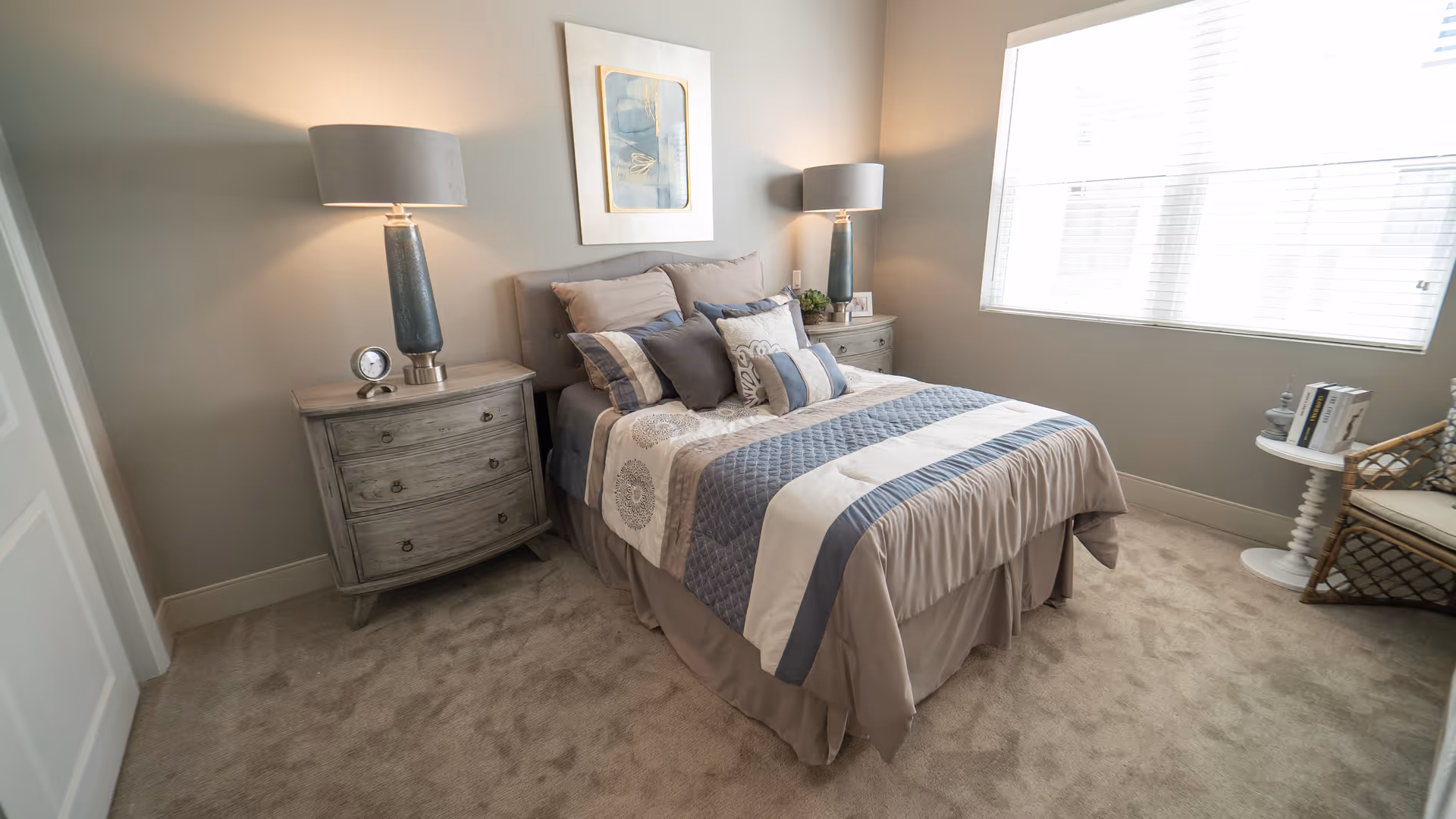A neatly made bed with multiple pillows in a cozy bedroom featuring two bedside tables with matching lamps, a framed artwork above the bed, a window with blinds allowing natural light, a small round table with books, and a wicker chair.