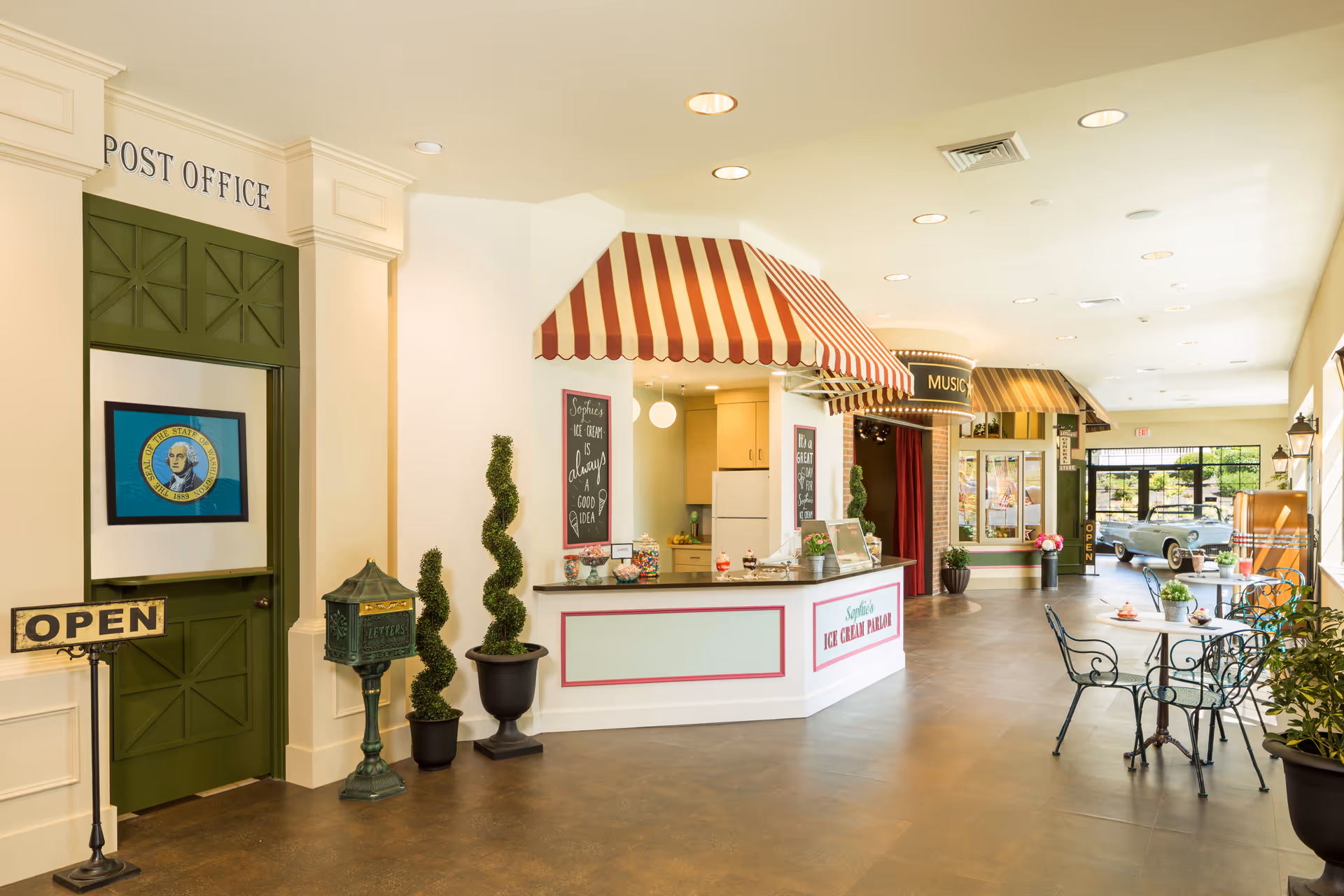 Bright indoor common area featuring a striped ice cream parlor counter, small café tables, and a 'Post Office' door.