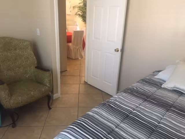A small bedroom with a striped bedspread on the bed and a green upholstered armchair. The door is open, showing a glimpse of a dining area with a table covered by a red tablecloth and white slipcovered chairs.