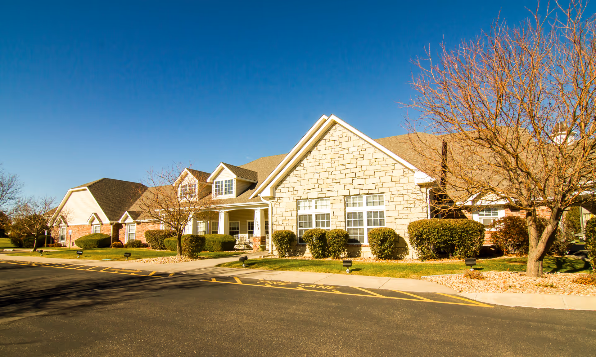 Front exterior of a single-story senior living building with a stone facade, shrubs, leafless trees, and a driveway under a clear blue sky.