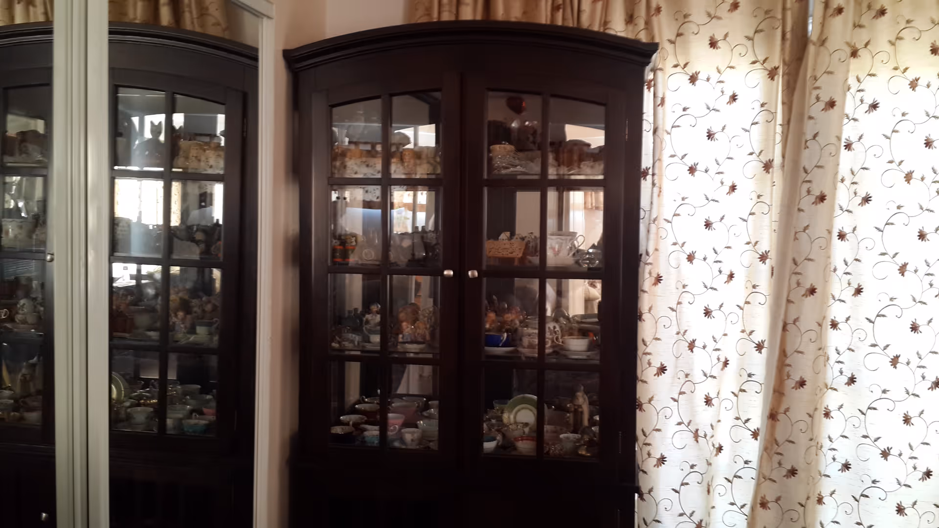 A dark wooden glass-front cabinet filled with various decorative items and dishes, positioned next to a window with cream-colored curtains featuring a floral pattern. The cabinet's reflection is visible in a nearby mirror.