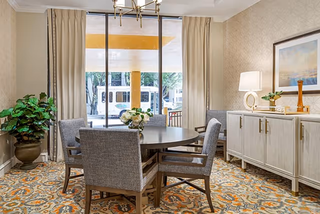 Bright dining room with a round table surrounded by upholstered chairs, a sideboard with a lamp and plant, and a large window looking out to the street.