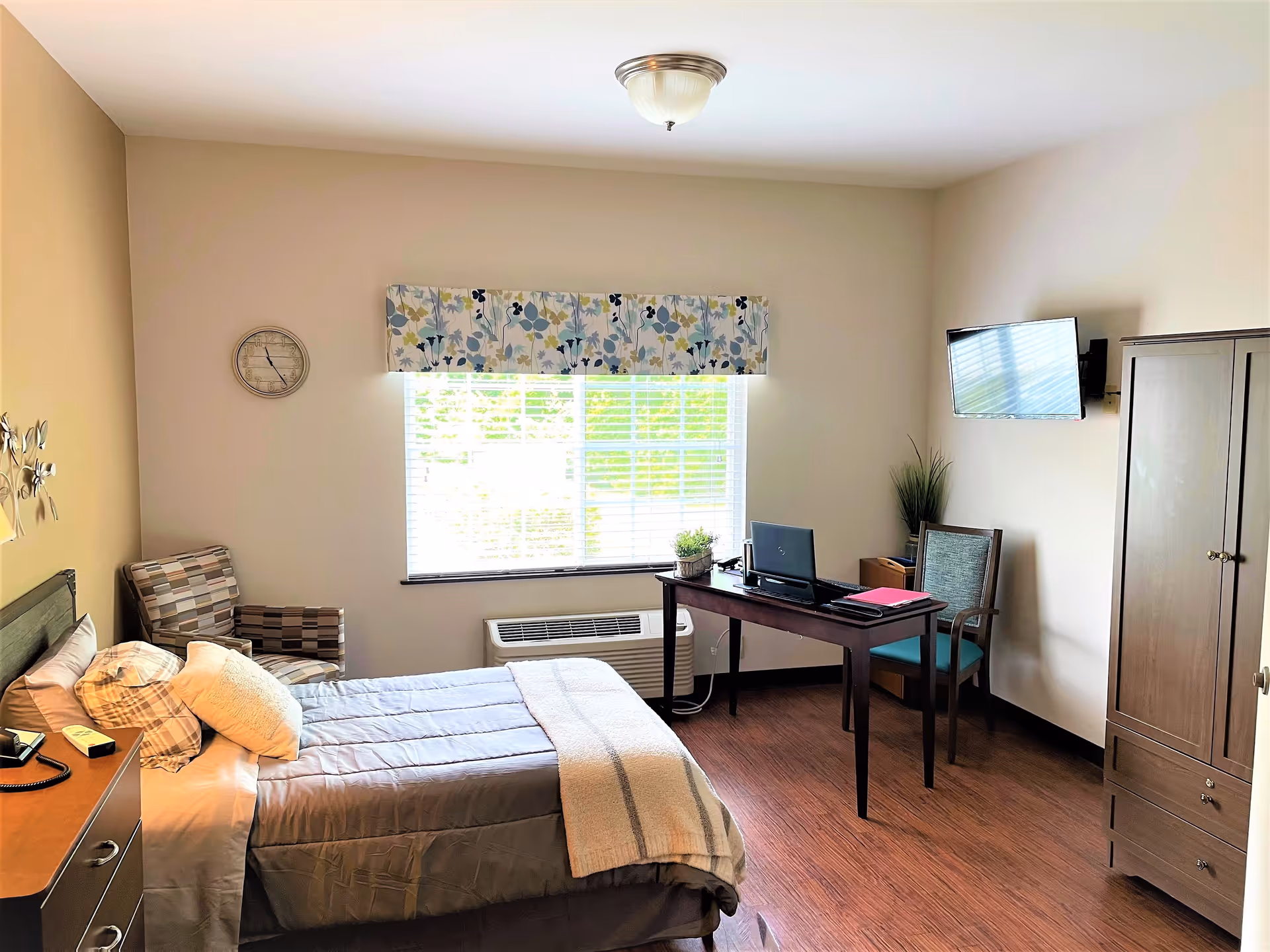 A bright and tidy bedroom in an assisted living facility featuring a single bed with pillows and a blanket, a wooden nightstand with a phone and clock, a patterned armchair, a large window with floral valance, a wooden desk with a laptop and chair, a wall-mounted TV, a tall wooden wardrobe, and wood flooring.