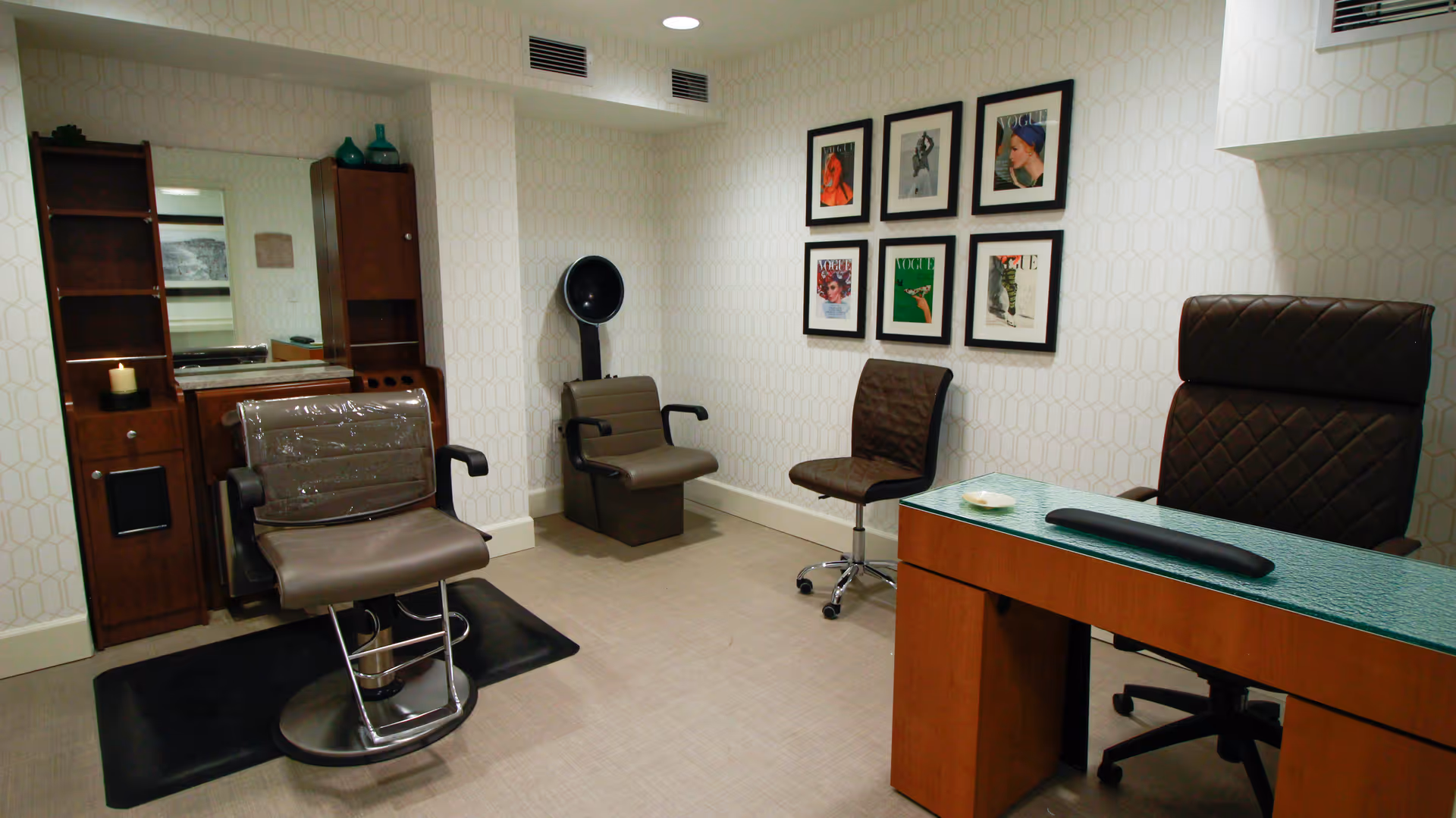 Interior of a salon room with a salon chair in front of a mirror and wooden cabinet, a hair dryer chair, a rolling chair, a desk with a glass top, and six framed Vogue magazine covers on the wall.
