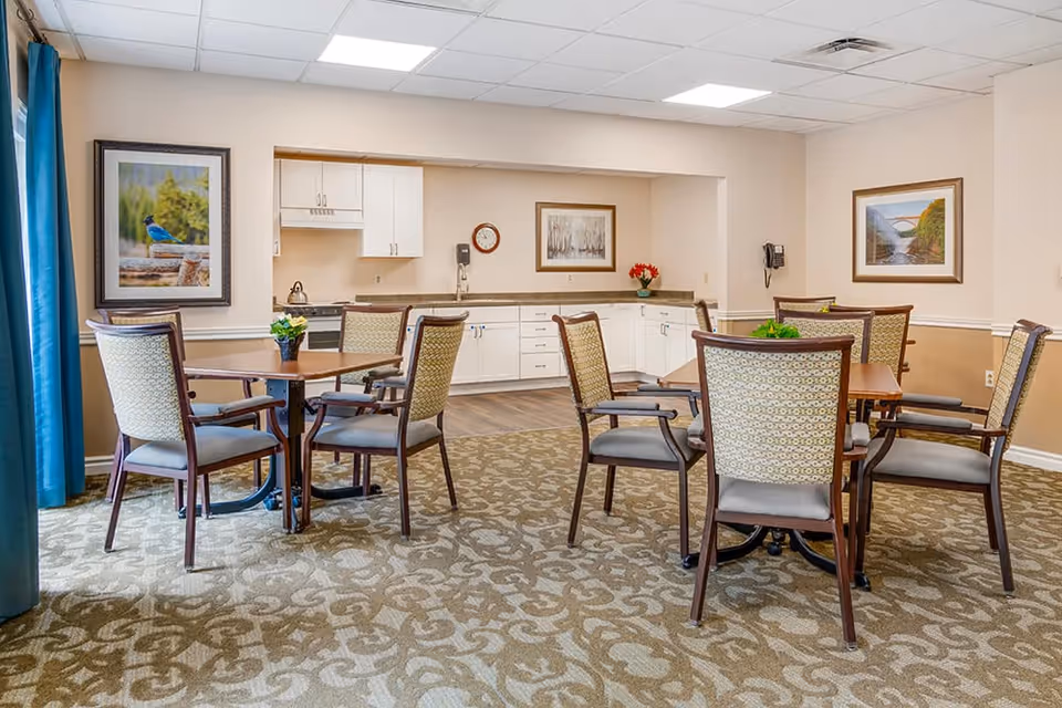 Communal dining area with several tables and upholstered chairs in front of a small kitchenette and framed artwork.