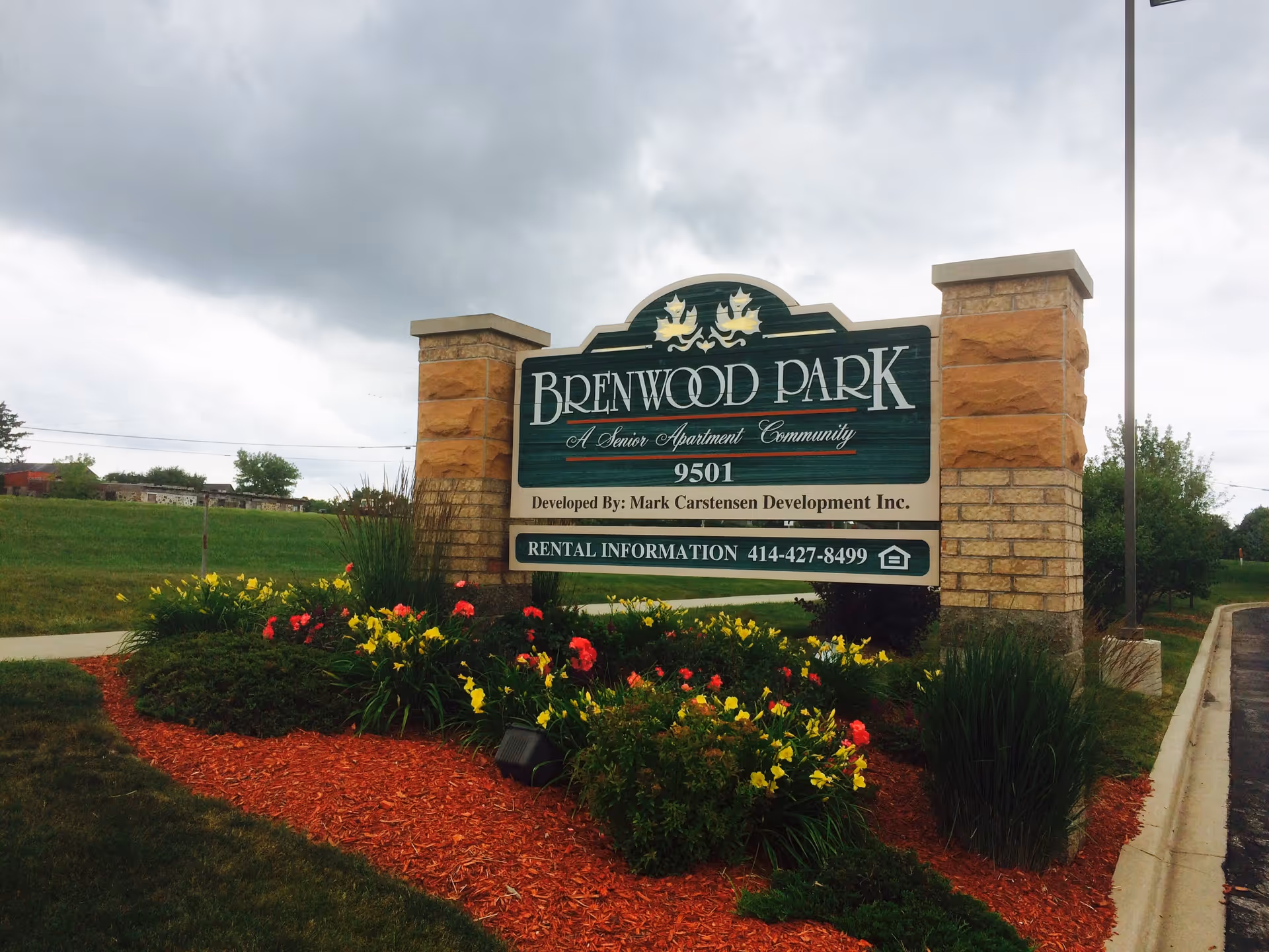 Entrance sign for Brenwood Park senior apartment community surrounded by landscaped flowers and mulch under a cloudy sky.
