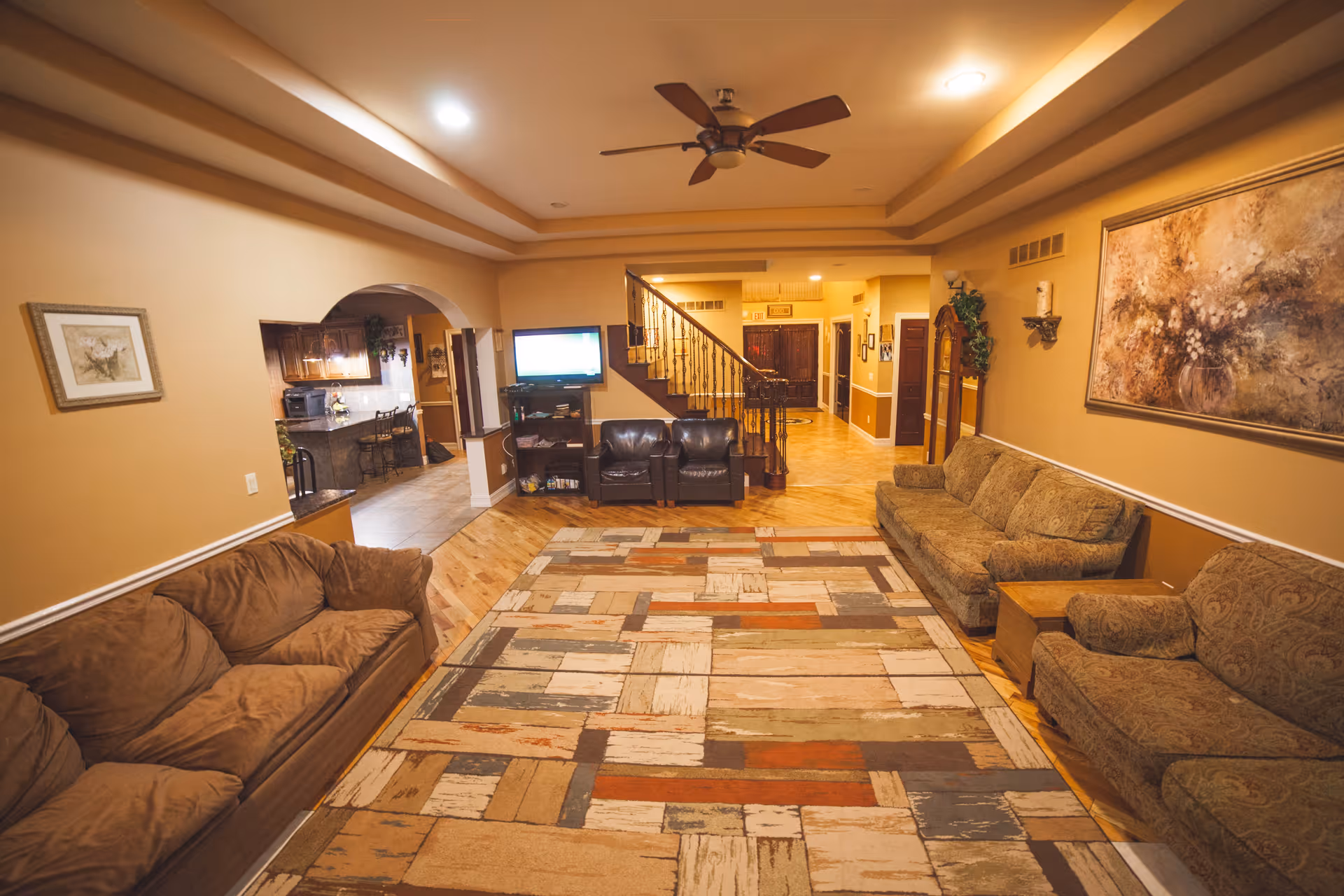 A spacious living room with three sofas arranged around a large patterned area rug. The room features warm beige walls, a ceiling fan, a large painting on the wall, and a staircase leading to an upper floor. In the background, there is a view into a kitchen area with bar stools and a television mounted on a stand near the stairs.