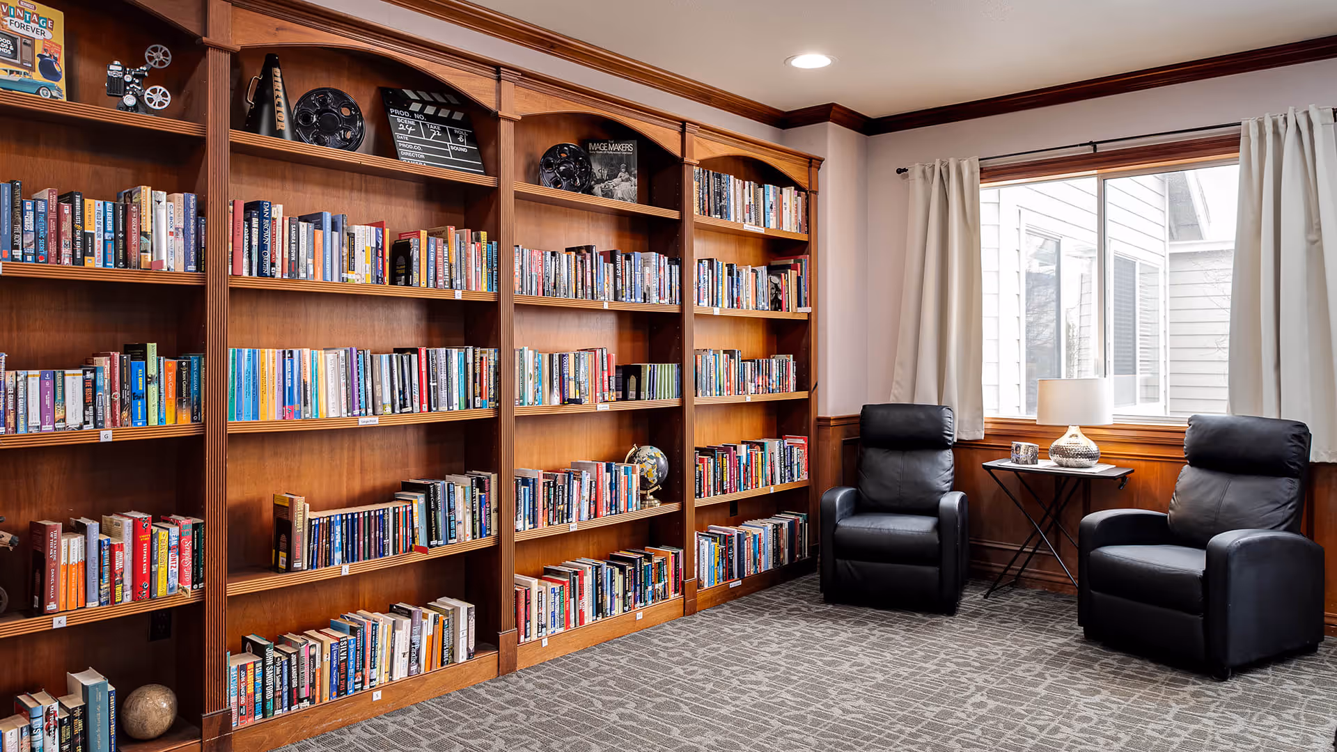 A cozy reading room with wooden bookshelves filled with books along one wall, two black leather recliners, a small side table with a lamp and framed photos, and a large window with white curtains letting in natural light.