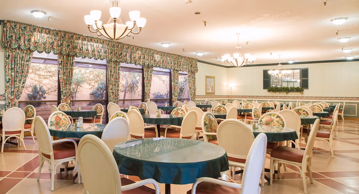 Spacious dining room with round tables covered in green tablecloths, floral-upholstered chairs, chandeliers, and large windows with floral curtains.