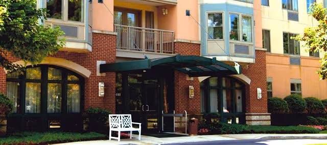 Entrance of a multistory senior living building with a brick facade, glass doors under a green canopy and a white bench out front.