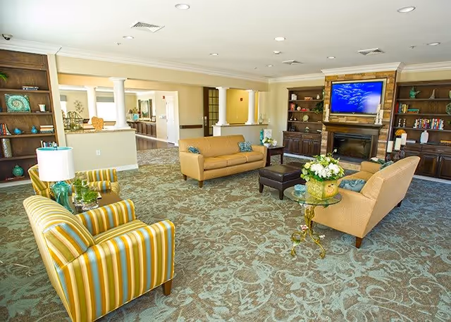 A spacious living room with patterned carpet, two striped armchairs, two beige sofas, a glass-top side table with a flower arrangement, and a dark ottoman. The room features built-in wooden shelves on either side of a fireplace with a mounted flat-screen TV above it. The walls are light-colored, and the ceiling has recessed lighting.