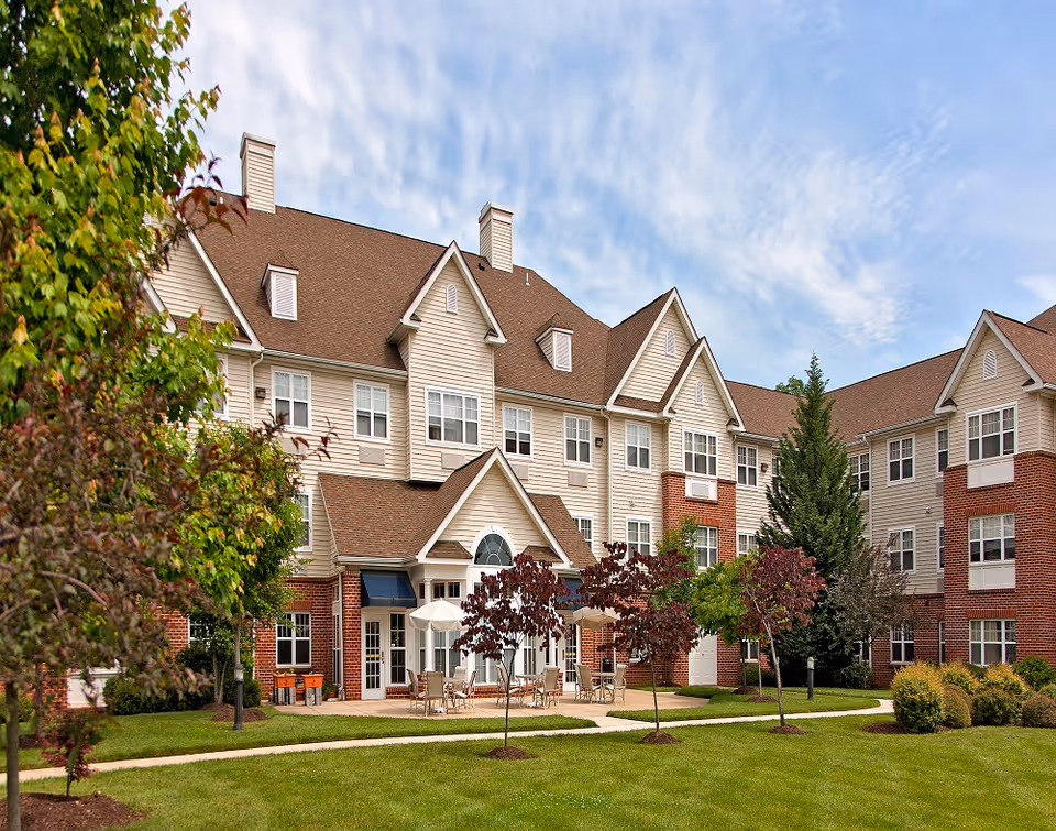 Exterior view of Hampshire Village senior living facility showing a multi-story building with beige siding and red brick accents. The building has multiple windows, peaked roofs, and chimneys. In front of the building is a well-maintained lawn with small trees, shrubs, and a paved walkway leading to a patio area with tables, chairs, and umbrellas under a partly cloudy sky.
