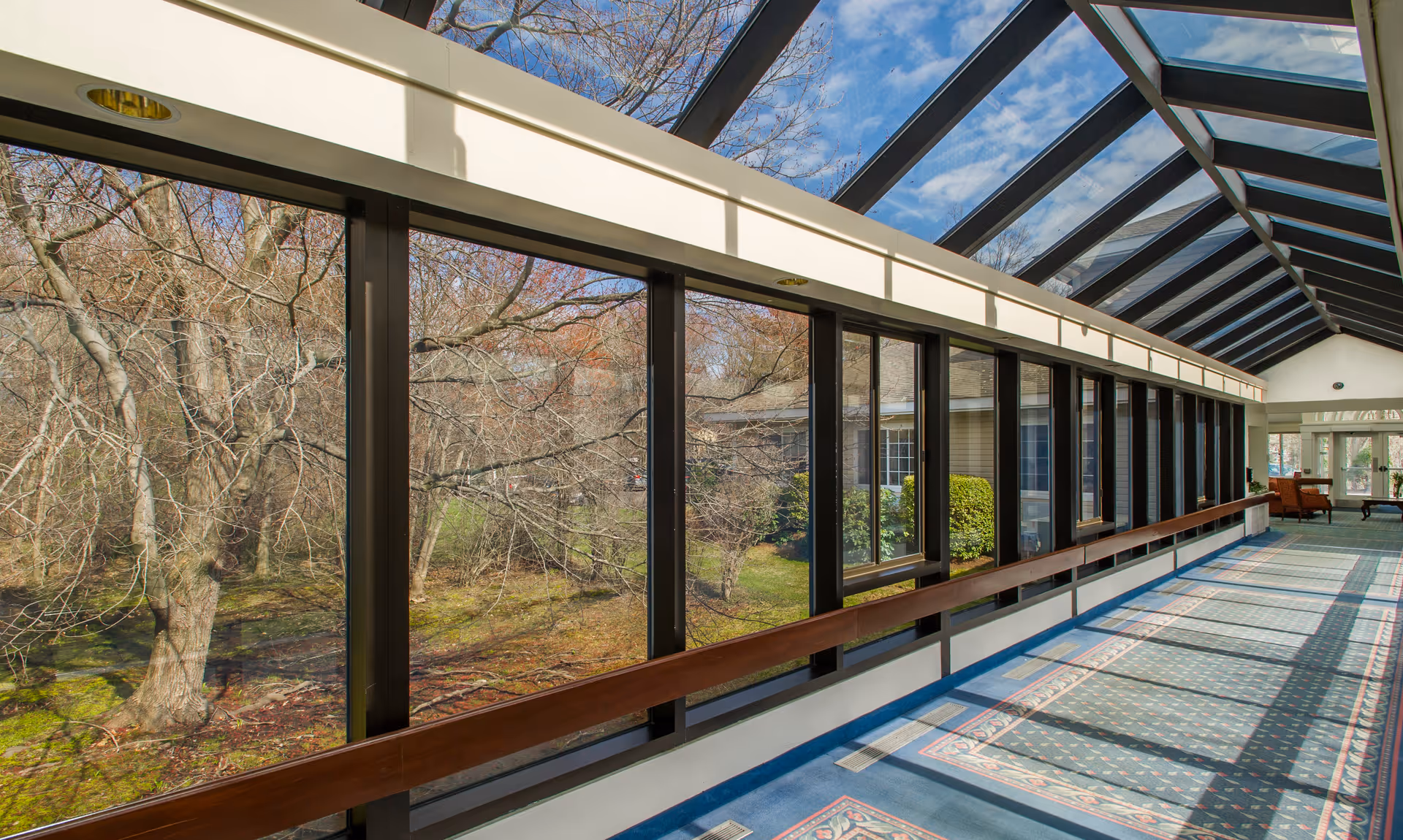A long indoor corridor with large floor-to-ceiling windows on one side and a glass ceiling allowing natural light to fill the space. Outside the windows, leafless trees and greenery are visible. The corridor has a blue carpet with patterned borders and a wooden handrail along the windows.