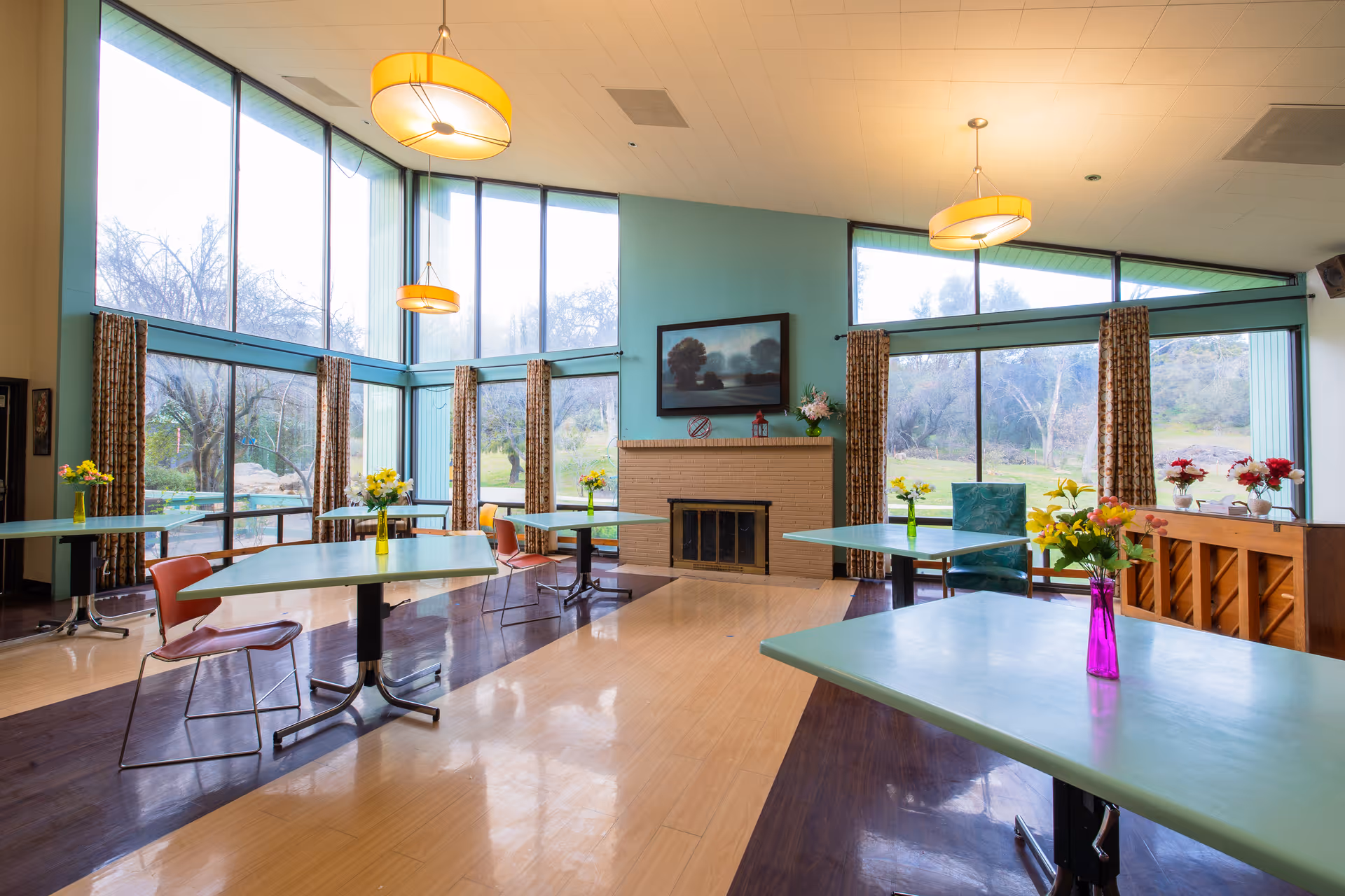 Bright communal dining/activity room with large windows, multiple tables and chairs, a fireplace, and vases of flowers.