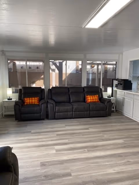 Bright common room with black leather reclining sofa and armchair in front of large windows, light wood flooring, side tables with lamps and a small kitchenette area.