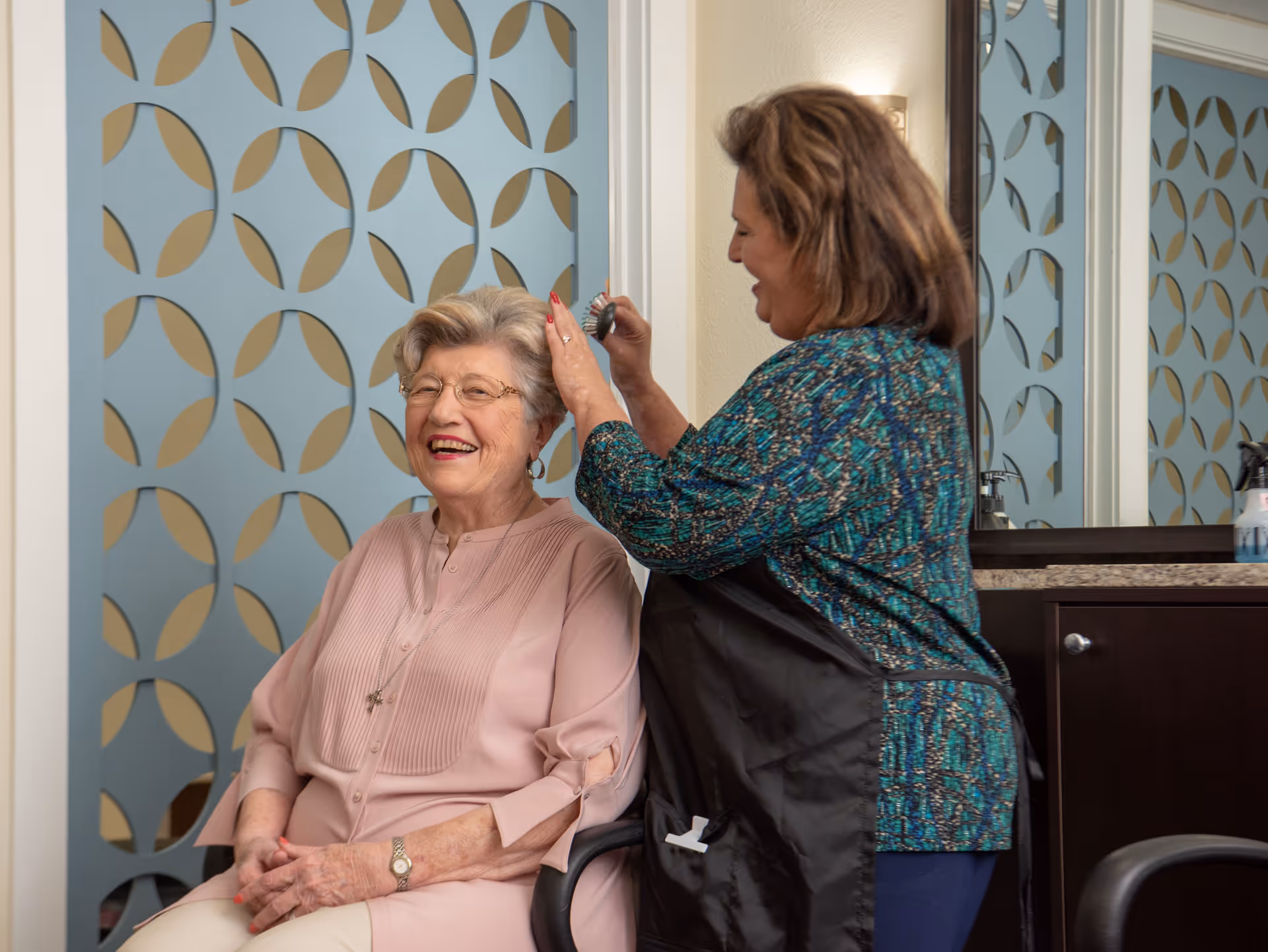 An elderly woman with glasses and a pink blouse is smiling while sitting in a chair as a hairstylist brushes her hair in a salon setting with decorative blue and beige patterned wall panels and a mirror in the background.