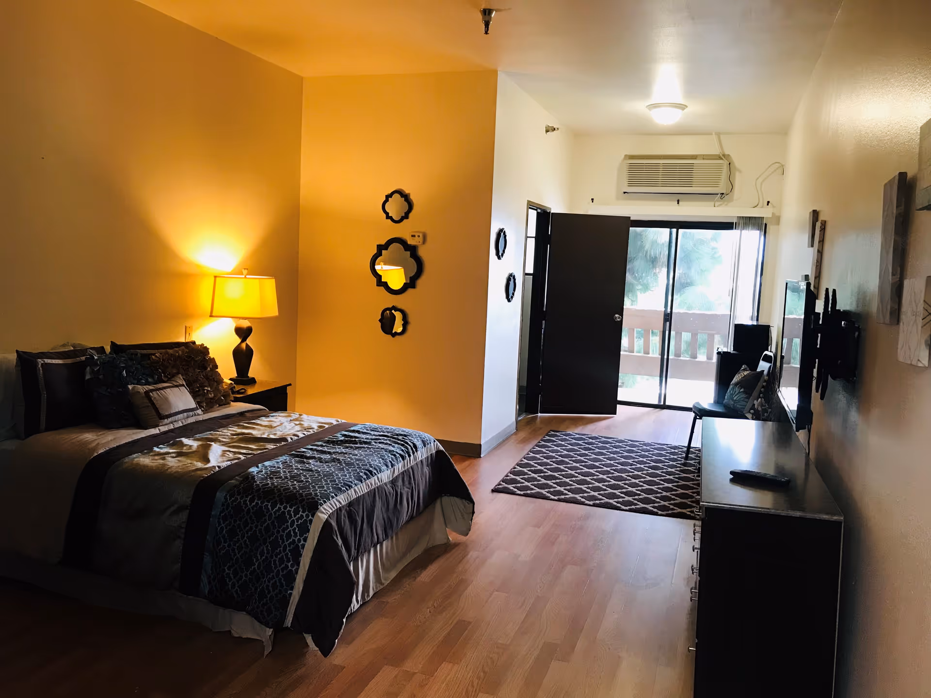 A cozy bedroom in a retirement center featuring a neatly made bed with decorative pillows and a bedside lamp emitting warm light. The room has wooden flooring, a dresser with a TV mounted on the wall above it, a chair with a cushion, and a sliding glass door leading to a balcony with a view of trees outside. Several decorative mirrors are hung on the wall near the bed.