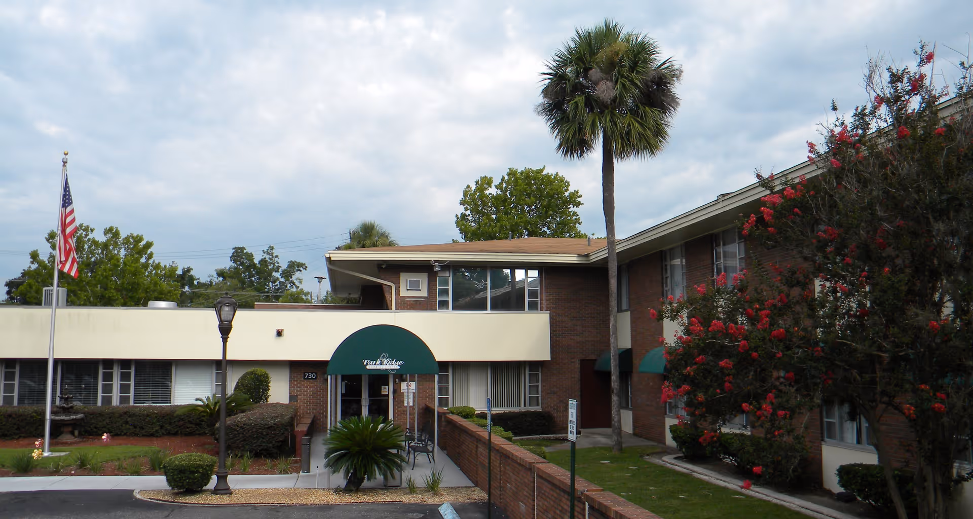 Front entrance of a two-story brick nursing and rehabilitation building with a green awning reading 'Park Ridge', a palm tree, American flag, and landscaped grounds.